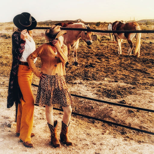 two women in western apparel, western hats western boots outside with horses in background