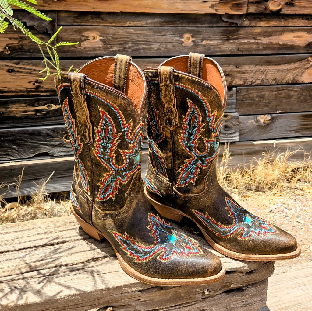 A pair of women's mid-calf leather boots, "Starbright" by Dan Post, in distressed brown with vibrant multi-colored stitching in a starburst design. The boots have a snip toe and low heel, placed on a black box with "Dan Post Handcrafted Cushion Comfort" branding, set against a rustic wooden wall and window backdrop.