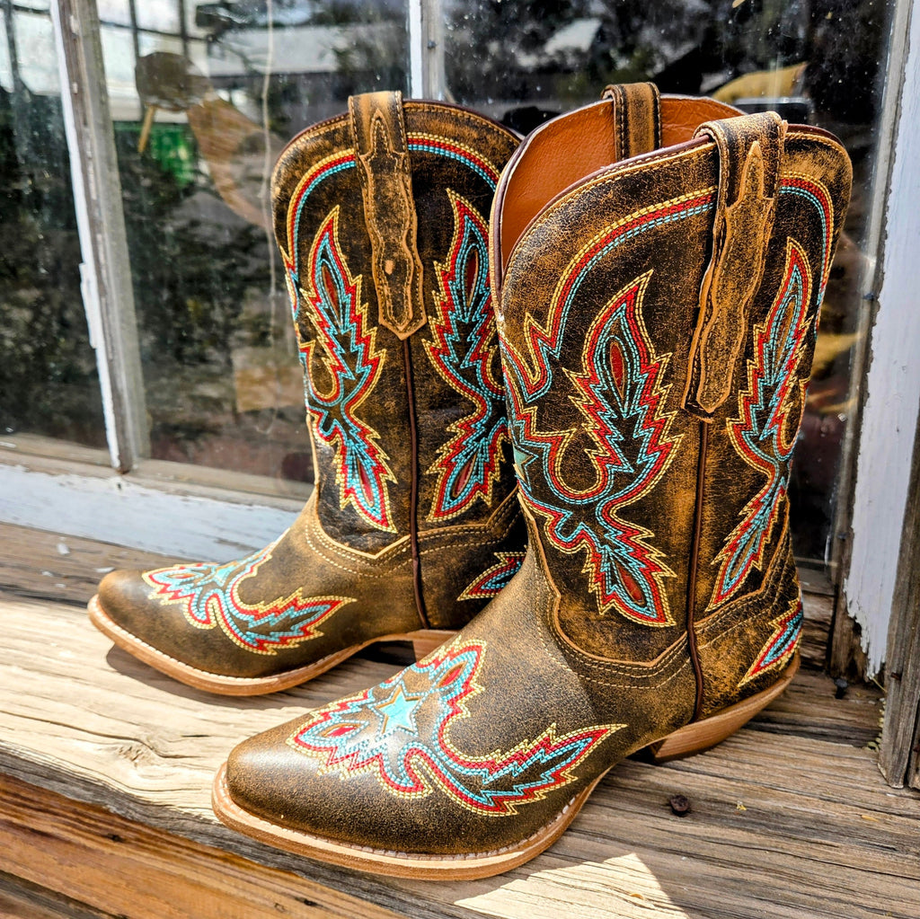A pair of women's mid-calf leather boots, "Starbright" by Dan Post, in distressed brown with vibrant multi-colored stitching in a starburst design. The boots have a snip toe and low heel, placed on a black box with "Dan Post Handcrafted Cushion Comfort" branding, set against a rustic wooden wall and window backdrop.