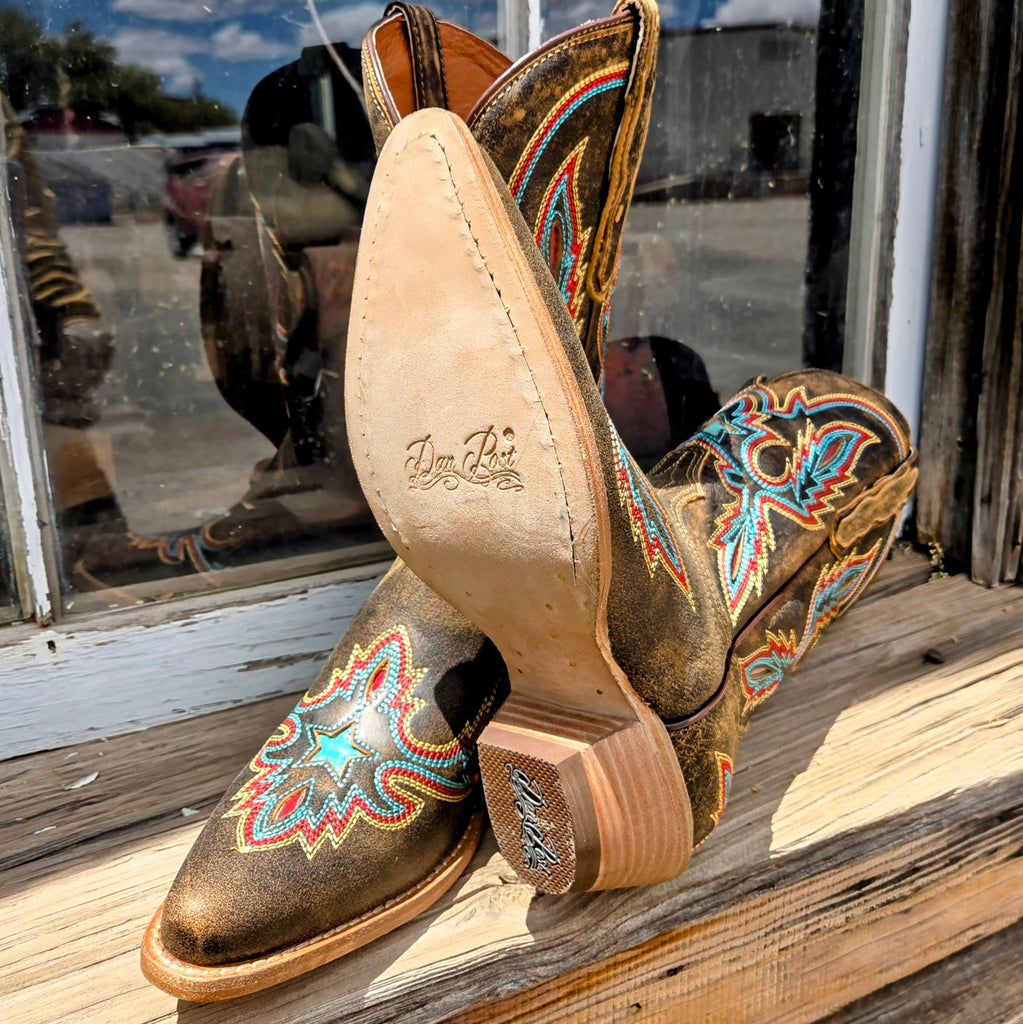 A pair of women's mid-calf leather boots, "Starbright" by Dan Post, in distressed brown with vibrant multi-colored stitching in a starburst design. The boots have a snip toe and low heel, placed on a black box with "Dan Post Handcrafted Cushion Comfort" branding, set against a rustic wooden wall and window backdrop.