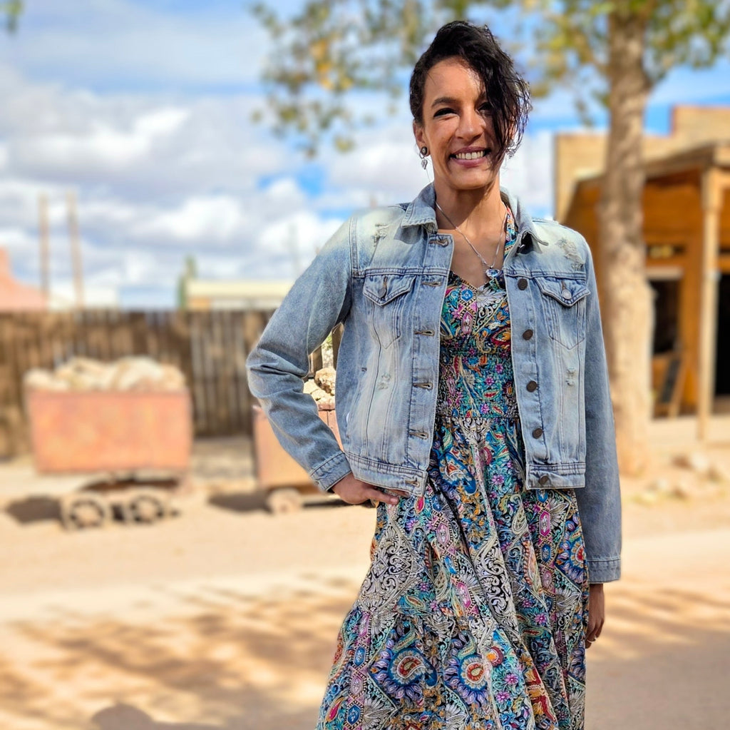 Stylish woman posing on a dusty street in historic Tombstone, Arizona, wearing a distressed light blue denim jacket featuring a vibrant bedazzled cowgirl boots design on the back by Forward Design