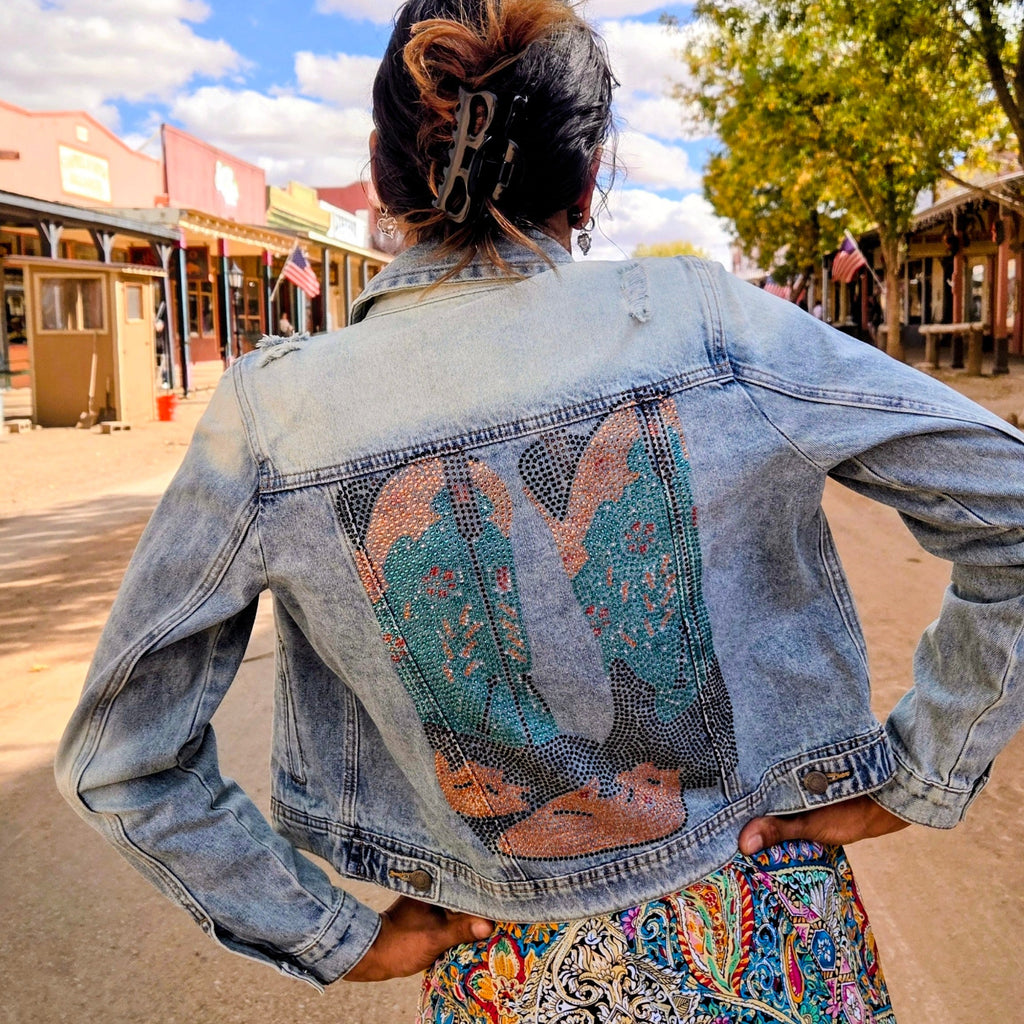 Stylish woman posing on a dusty street in historic Tombstone, Arizona, wearing a distressed light blue denim jacket featuring a vibrant bedazzled cowgirl boots design on the back by Forward Design