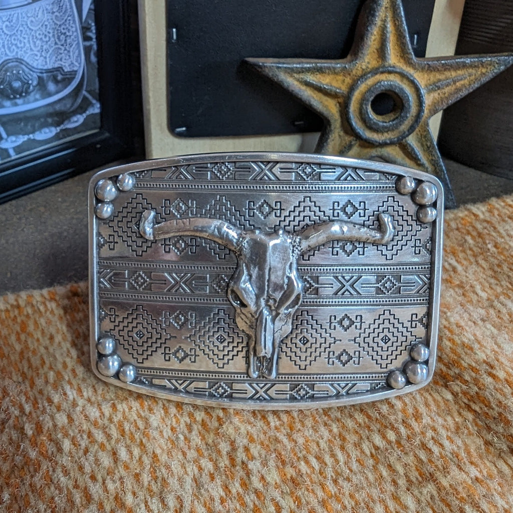 A belt buckle with a longhorn skull, framed by aztec patterns.