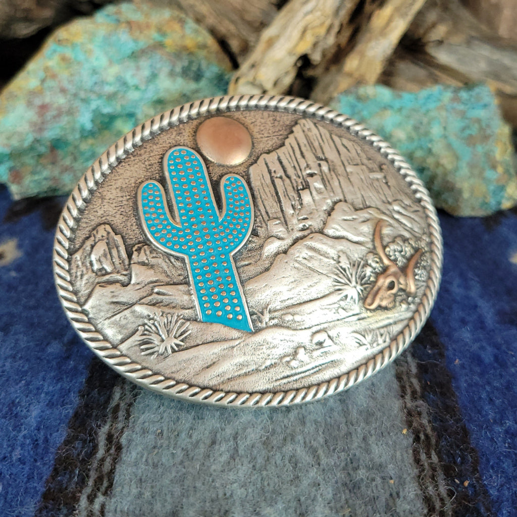 A belt buckle showcases a desert scene with a blue cactus, copper moon and skull, and etched mountains.