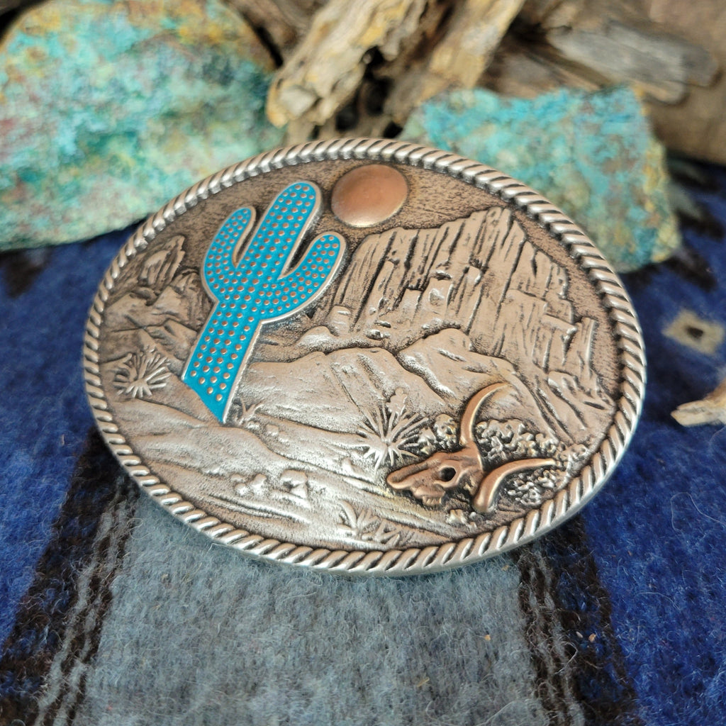 A belt buckle showcases a desert scene with a blue cactus, copper moon and skull, and etched mountains.