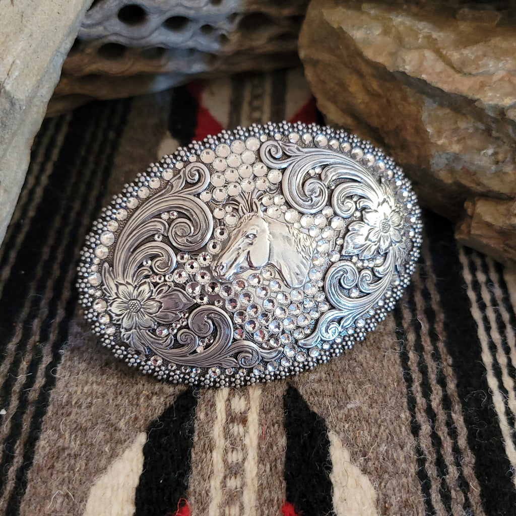 A belt buckle with a crystal-embellished horse head, framed by tooled silver.