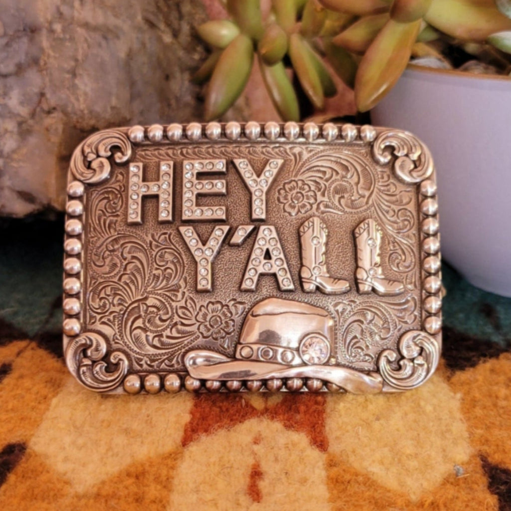 A belt buckle with "Hey Y'all" in rhinestone letters, featuring a cowboy hat.