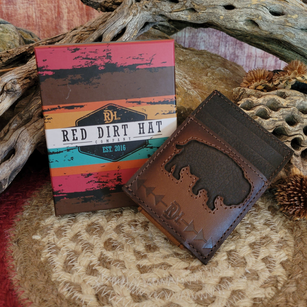 A brown leather Bison Money Clip Wallet by Red Dirt Hat Co. sits on a woven mat, next to its colorful packaging box featuring the Red Dirt Hat Co. logo and text "Est. 2016." The wallet showcases a laser-etched bison silhouette with arrowhead stitching, set against a rustic backdrop of driftwood and dried desert plants, capturing a true Western aesthetic.