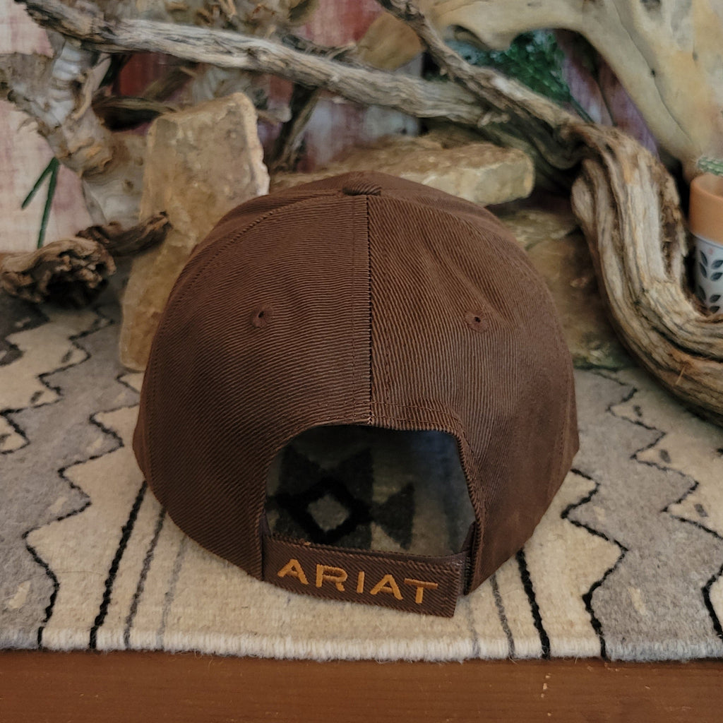 Brown trucker cap by Ariat with a black mesh back, featuring a curved bill with barbed wire stitching and an Ariat logo patch on the front. Displayed on a woven mat with a geometric pattern, set against a rustic backdrop of stacked rocks and driftwood. Back View