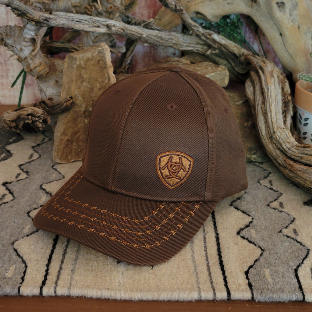 Brown trucker cap by Ariat with a black mesh back, featuring a curved bill with barbed wire stitching and an Ariat logo patch on the front. Displayed on a woven mat with a geometric pattern, set against a rustic backdrop of stacked rocks and driftwood. Front Side View
