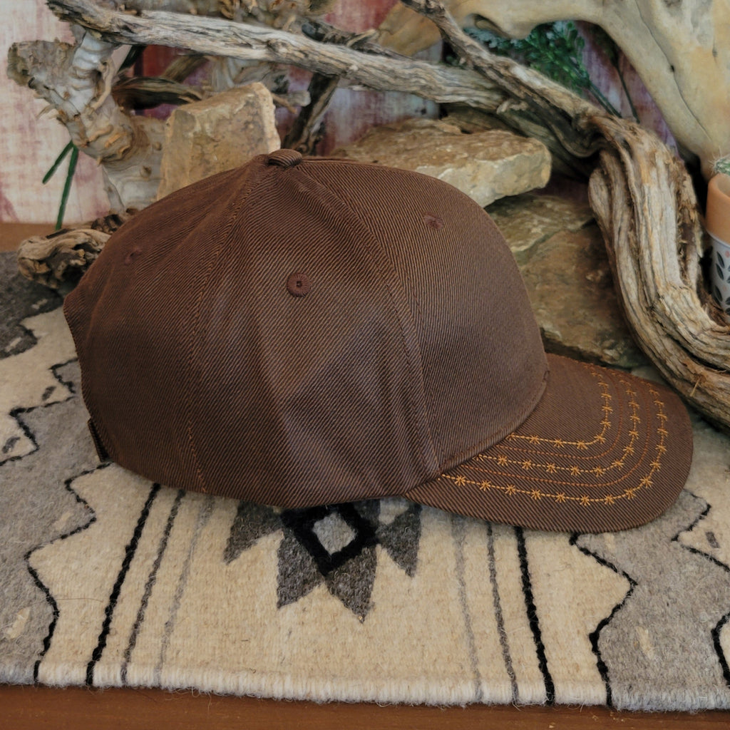 Brown trucker cap by Ariat with a black mesh back, featuring a curved bill with barbed wire stitching and an Ariat logo patch on the front. Displayed on a woven mat with a geometric pattern, set against a rustic backdrop of stacked rocks and driftwood. Side View2