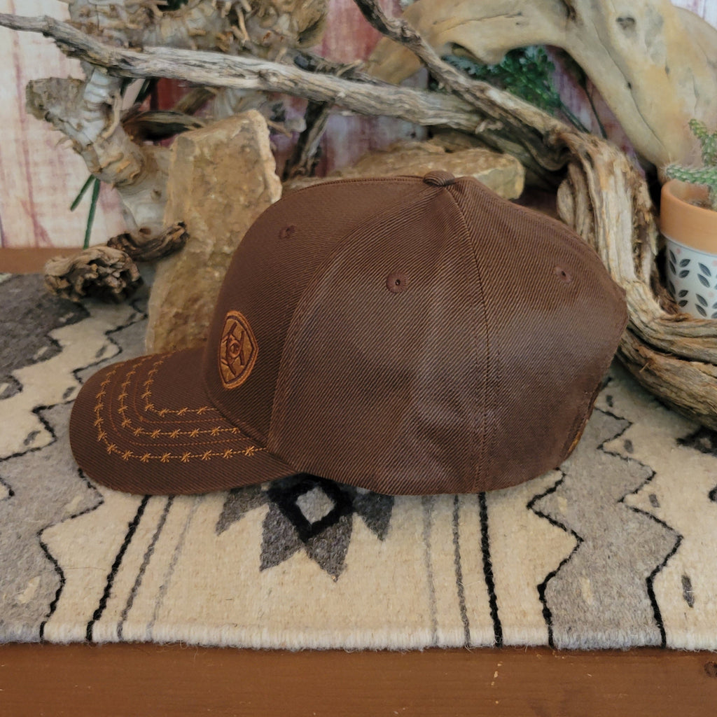 Brown trucker cap by Ariat with a black mesh back, featuring a curved bill with barbed wire stitching and an Ariat logo patch on the front. Displayed on a woven mat with a geometric pattern, set against a rustic backdrop of stacked rocks and driftwood. Side View