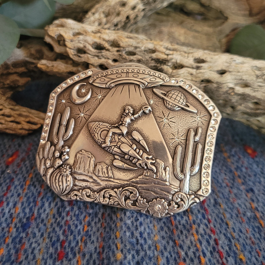 Silver belt buckle featuring a cosmic desert scene with a UFO, cowboy, and cacti, accented with studs.