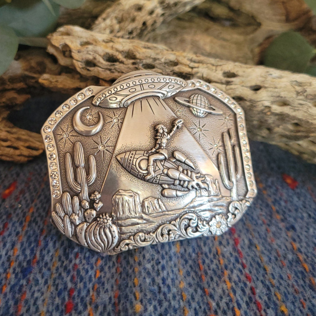 Silver belt buckle featuring a cosmic desert scene with a UFO, cowboy, and cacti, accented with studs.