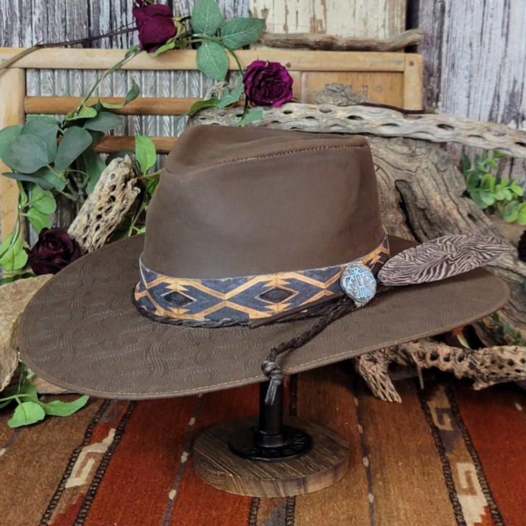A brown leather hat with a diamond-patterned band, featuring a turquoise Thunderbird concho and large patterned accent.