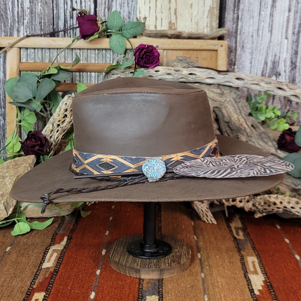 A brown leather hat with a diamond-patterned band, featuring a turquoise Thunderbird concho and large patterned accent.