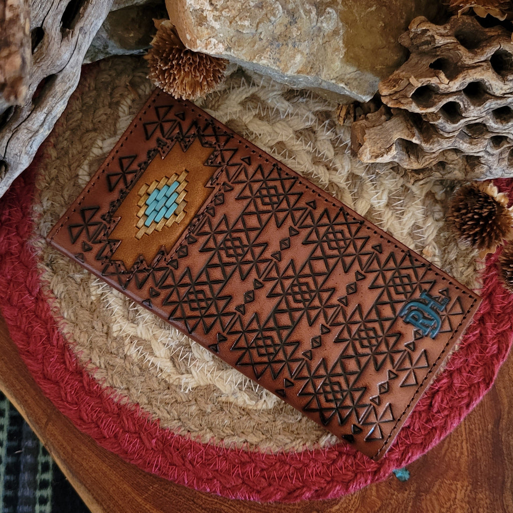 A brown leather Men's Laced Southwestern Medallion Rodeo Wallet by Red Dirt Hat Co. rests on a woven mat with a red border, surrounded by rustic driftwood and dried desert plants. The wallet features a turquoise and tan medallion with laced detailing and geometric cutouts, with the Red Dirt Hat Co. logo embossed, set against a natural Western backdrop.