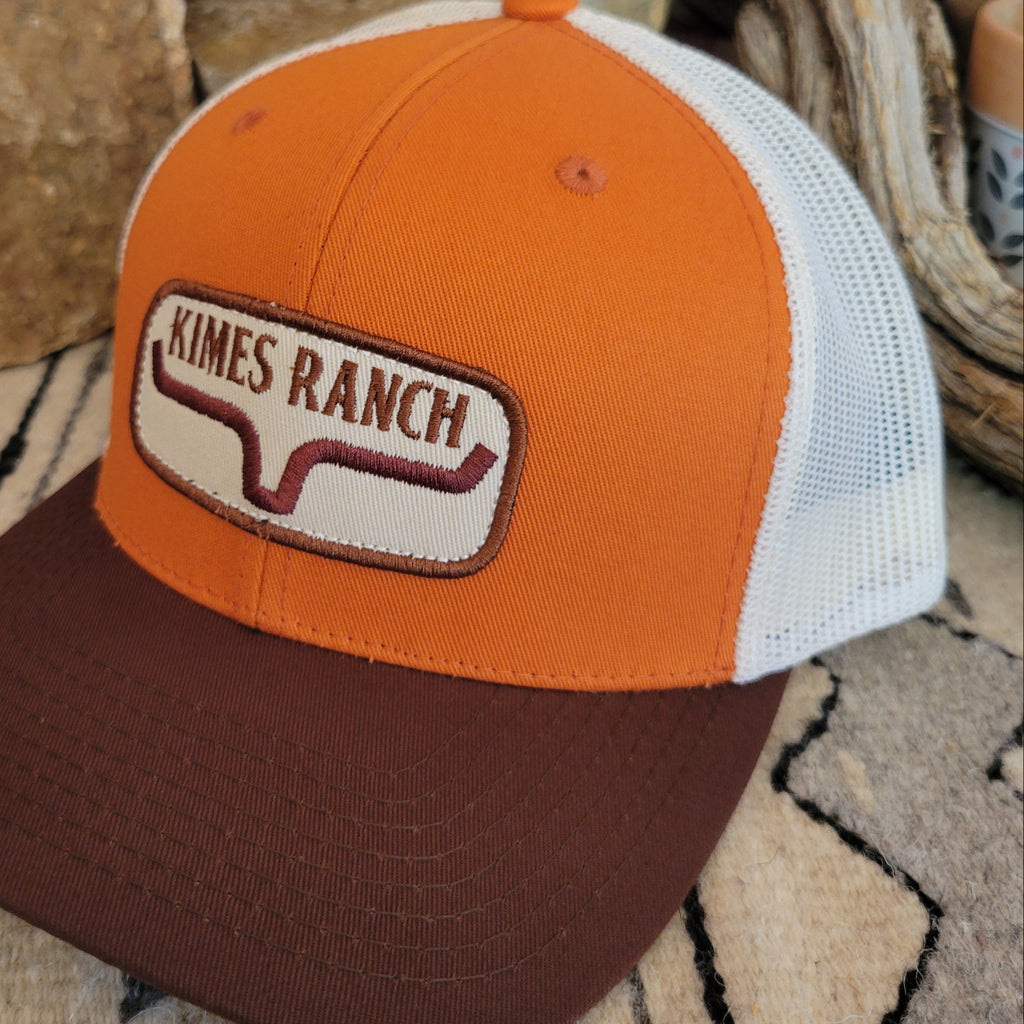 Orange trucker cap by Kimes Ranch with a brown bill and white mesh back, featuring an embroidered “Kimes Ranch” patch with a cattle brand design. Displayed on a woven mat with a geometric pattern, set against a rustic backdrop of stacked rocks, driftwood, and a potted succulent. Detailed View