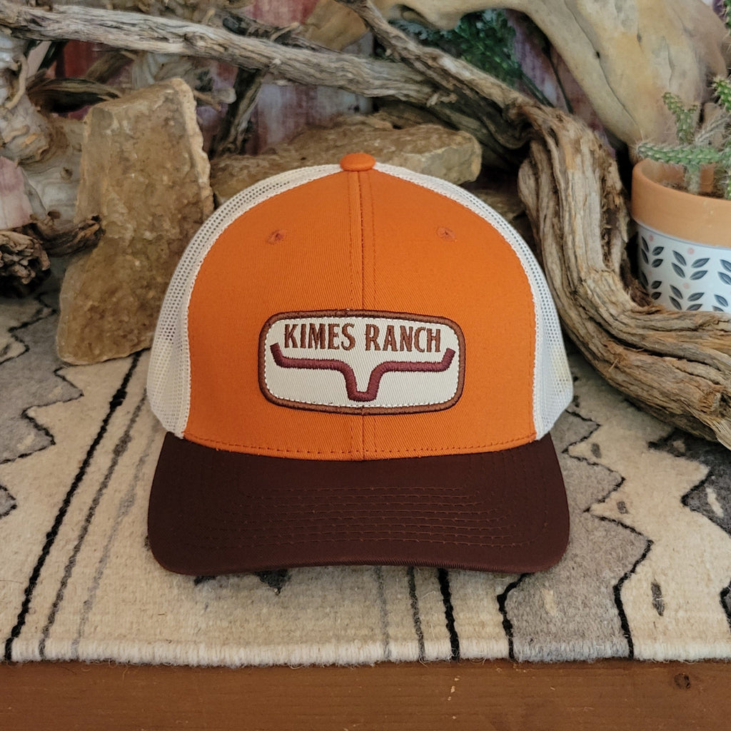 Orange trucker cap by Kimes Ranch with a brown bill and white mesh back, featuring an embroidered “Kimes Ranch” patch with a cattle brand design. Displayed on a woven mat with a geometric pattern, set against a rustic backdrop of stacked rocks, driftwood, and a potted succulent. Front View