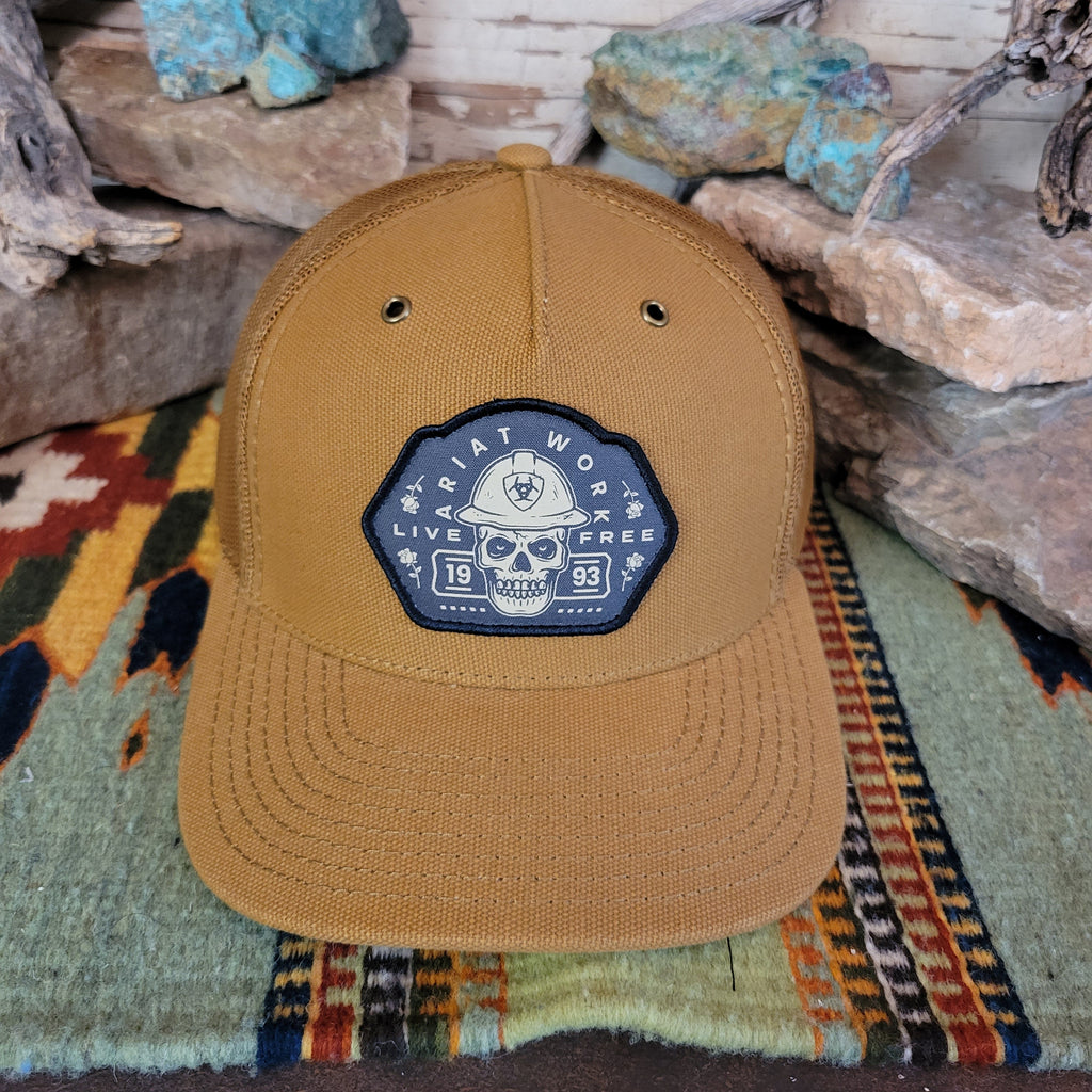 A tan trucker cap with a patch featuring a skull patch with text.