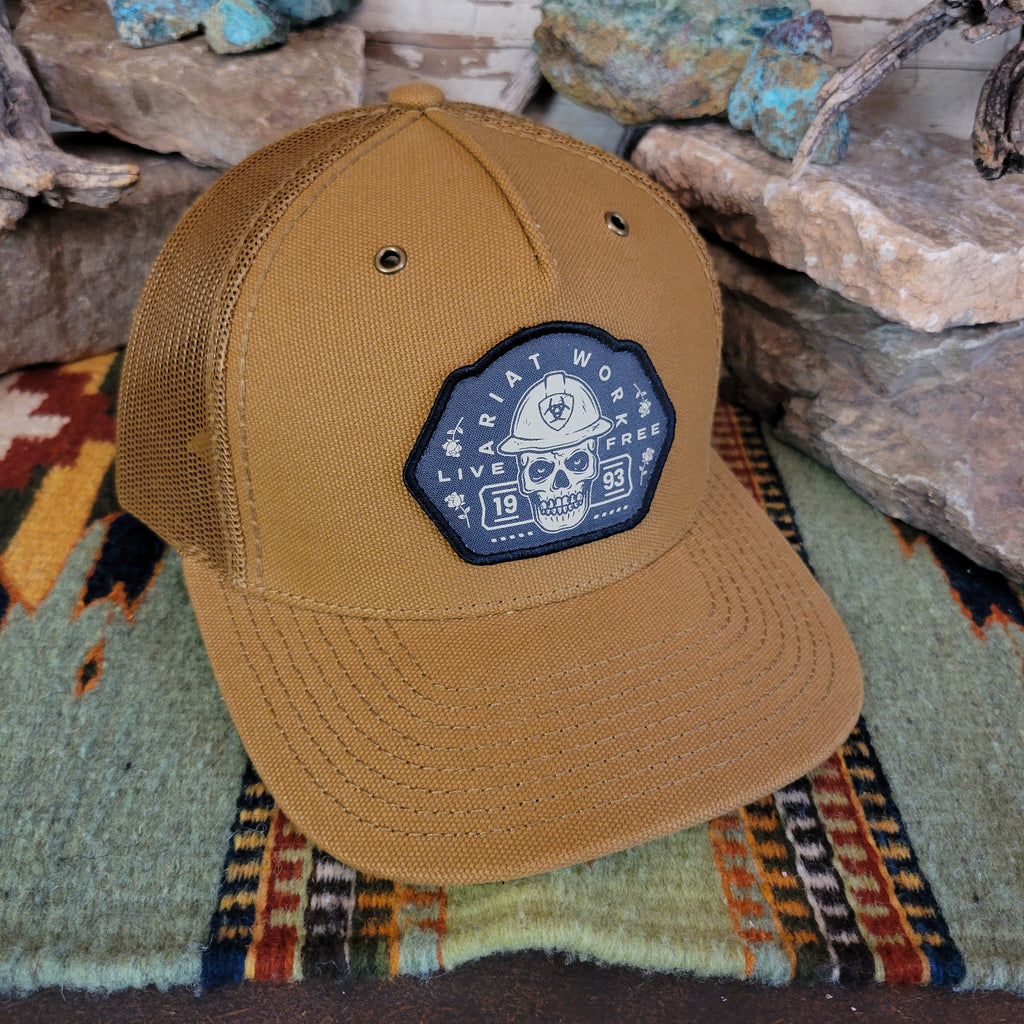 A tan trucker cap with a patch featuring a skull patch with text.