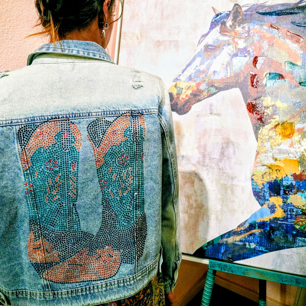 Stylish woman posing on a dusty street in historic Tombstone, Arizona, wearing a distressed light blue denim jacket featuring a vibrant bedazzled cowgirl boots design on the back by Forward Design