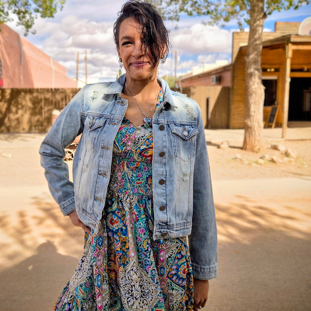 Stylish woman posing on a dusty street in historic Tombstone, Arizona, wearing a distressed light blue denim jacket featuring a vibrant bedazzled cowgirl boots design on the back by Forward Design
