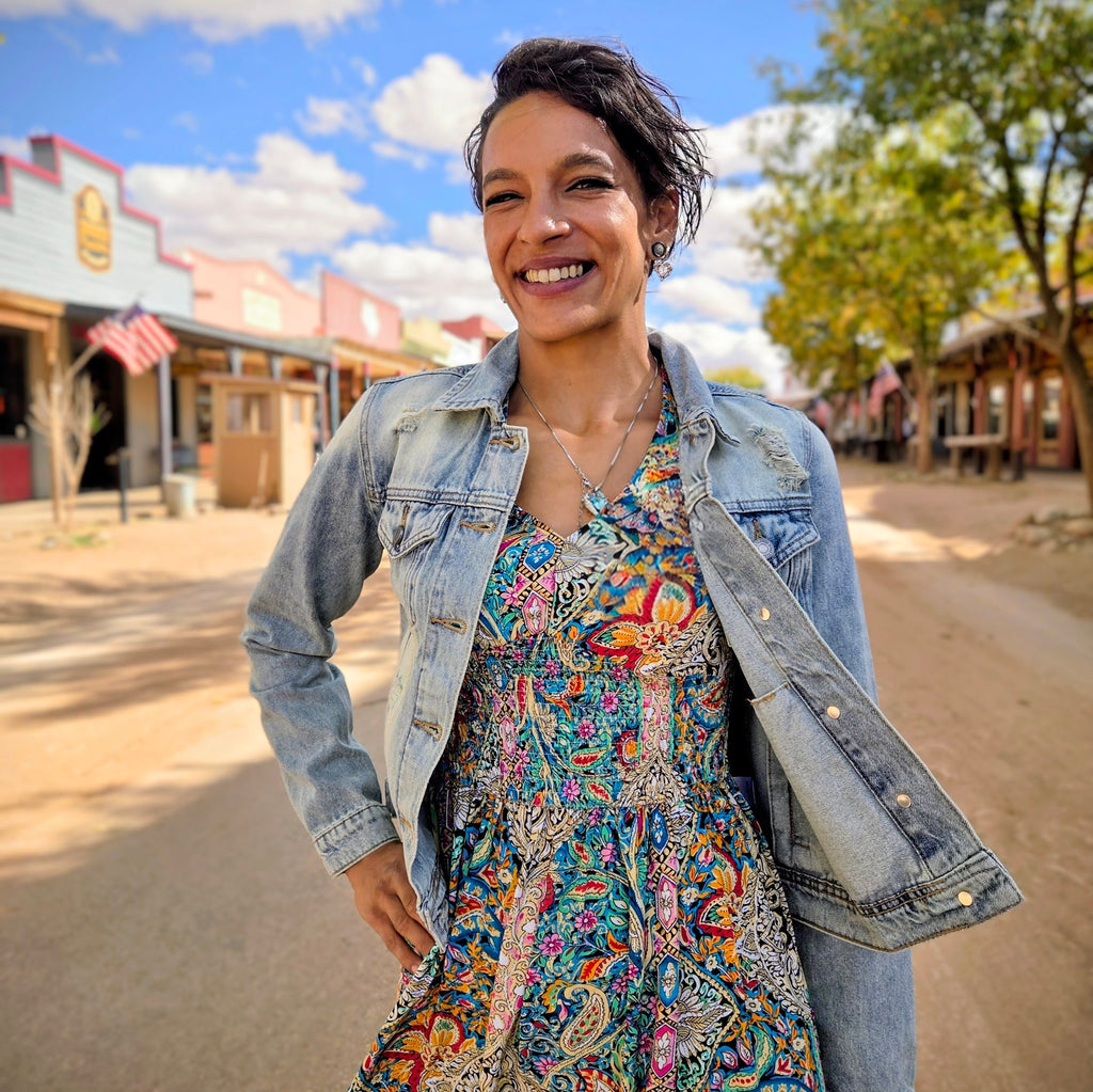 Stylish woman posing on a dusty street in historic Tombstone, Arizona, wearing a distressed light blue denim jacket featuring a vibrant bedazzled cowgirl boots design on the back by Forward Design