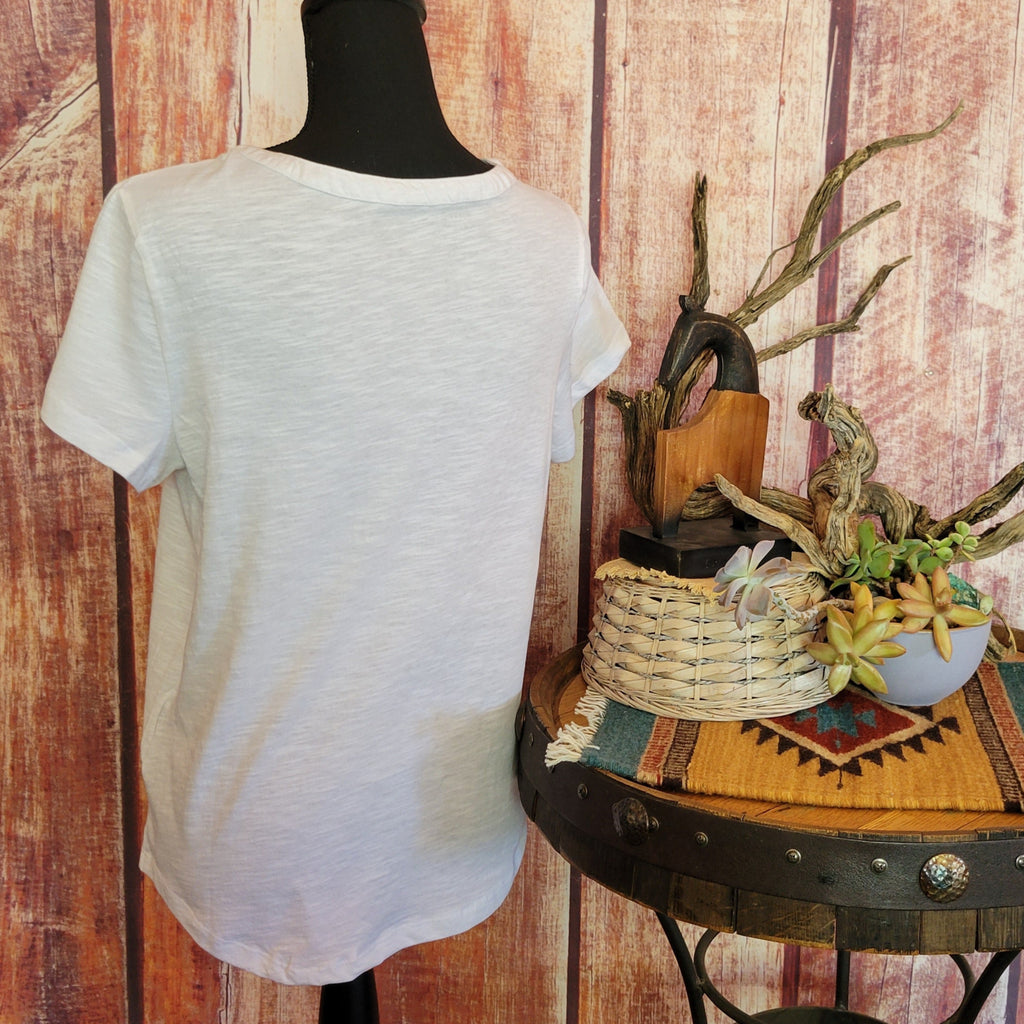 Women’s white knit V-neck shirt by Roper with short sleeves, featuring colorful floral embroidery in red, blue, and yellow across the chest. Displayed on a mannequin against a rustic wooden wall, with a barrel table holding a basket and potted succulents, accented by driftwood and a woven mat.Back View
