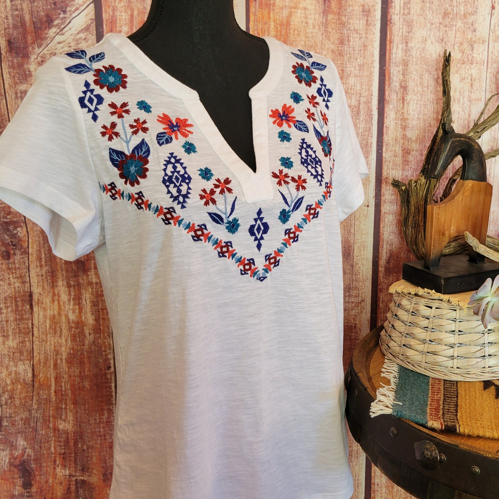 Women’s white knit V-neck shirt by Roper with short sleeves, featuring colorful floral embroidery in red, blue, and yellow across the chest. Displayed on a mannequin against a rustic wooden wall, with a barrel table holding a basket and potted succulents, accented by driftwood and a woven mat. Detailed Front View