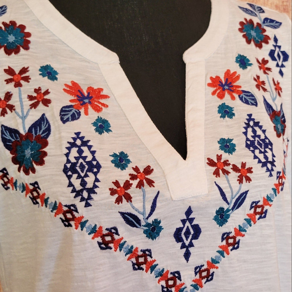 Women’s white knit V-neck shirt by Roper with short sleeves, featuring colorful floral embroidery in red, blue, and yellow across the chest. Displayed on a mannequin against a rustic wooden wall, with a barrel table holding a basket and potted succulents, accented by driftwood and a woven mat. Detailed View