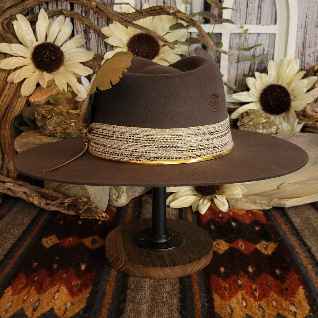 Brown cowgirl hat with decorative tan band, gold cords and feather, and a horseshoe stamp on the back.