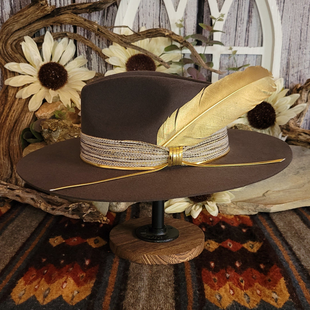Brown cowgirl hat with decorative tan band, gold feather and cords.