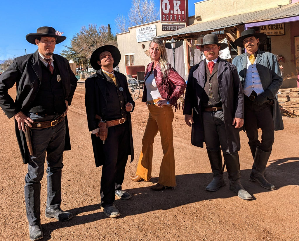 Female model wearing mustard colored western pants and  western boots with a western style red leather jacket surrounded by men dressed in old west attire, like Wyatt Earp & Doc Holliday. They are standing on historic Allen Street in front of the Ok Corral.