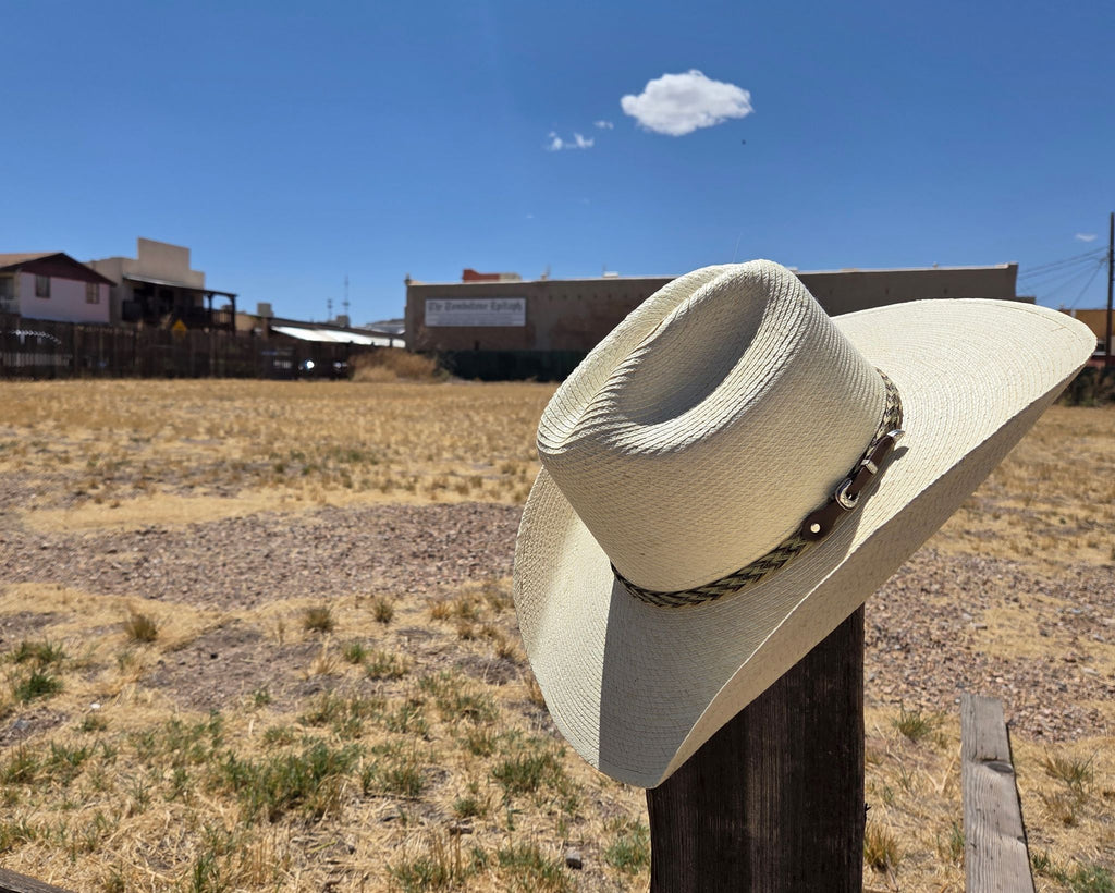  Image is of a creamy white palm straw hat, The Brooks by Larry Mahan, with a leather sweatband and branded logo. Wide brim visible, set against a dusty Tombstone backdrop under a clear blue sky with wooden fence and rustic buildings in the background.
