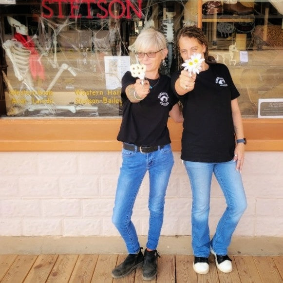 Two women in black 'You're a Daisy If You Do' T-shirts by Black Anchor Supply with daisy revolver graphic on back.