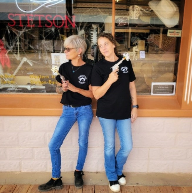 Two women in black 'You're a Daisy If You Do' T-shirts by Black Anchor Supply with daisy revolver graphic on back.