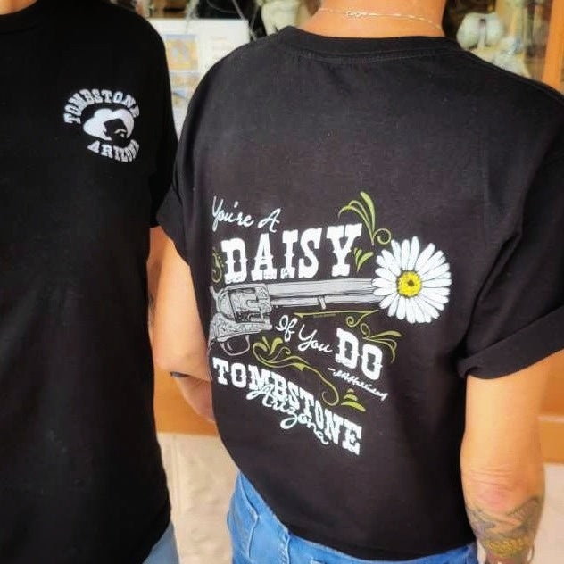Two women in black 'You're a Daisy If You Do' T-shirts by Black Anchor Supply with daisy revolver graphic on back.