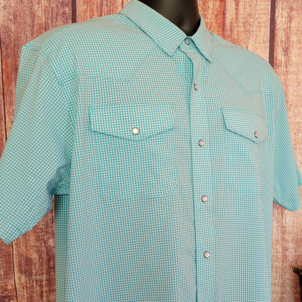 Men’s light turquoise checkered short-sleeve shirt by Ariat with snap buttons and two chest pockets, displayed on a mannequin against a rustic wooden wall. A barrel table nearby holds a basket and potted succulents, accented by driftwood, a bull skull sculpture, and a woven mat. Detail view.