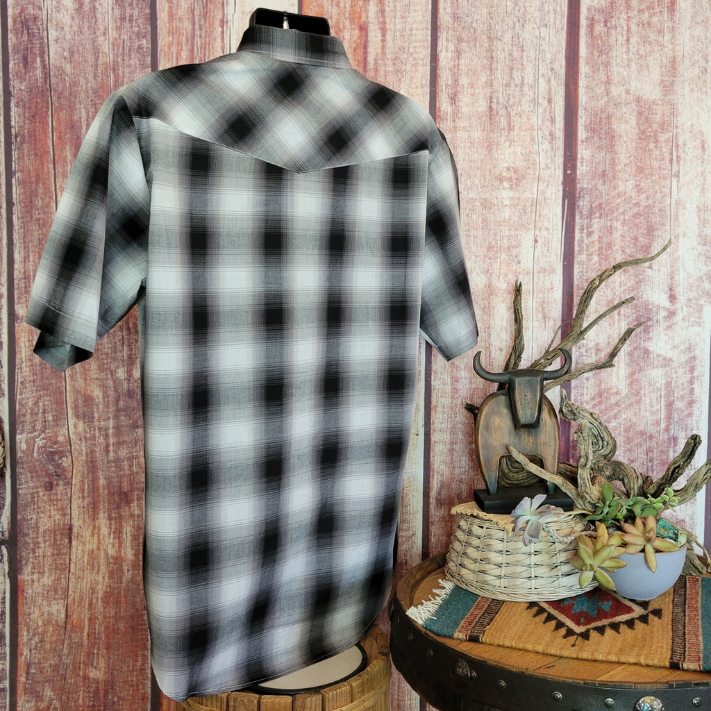 Men’s black plaid short-sleeve snap shirt by Roper on a mannequin, featuring white and gray accents and snap-button front. Set against a rustic wooden wall with a bull skull sculpture, dried branches, and a basket with succulents on a barrel table, adorned with a woven mat.