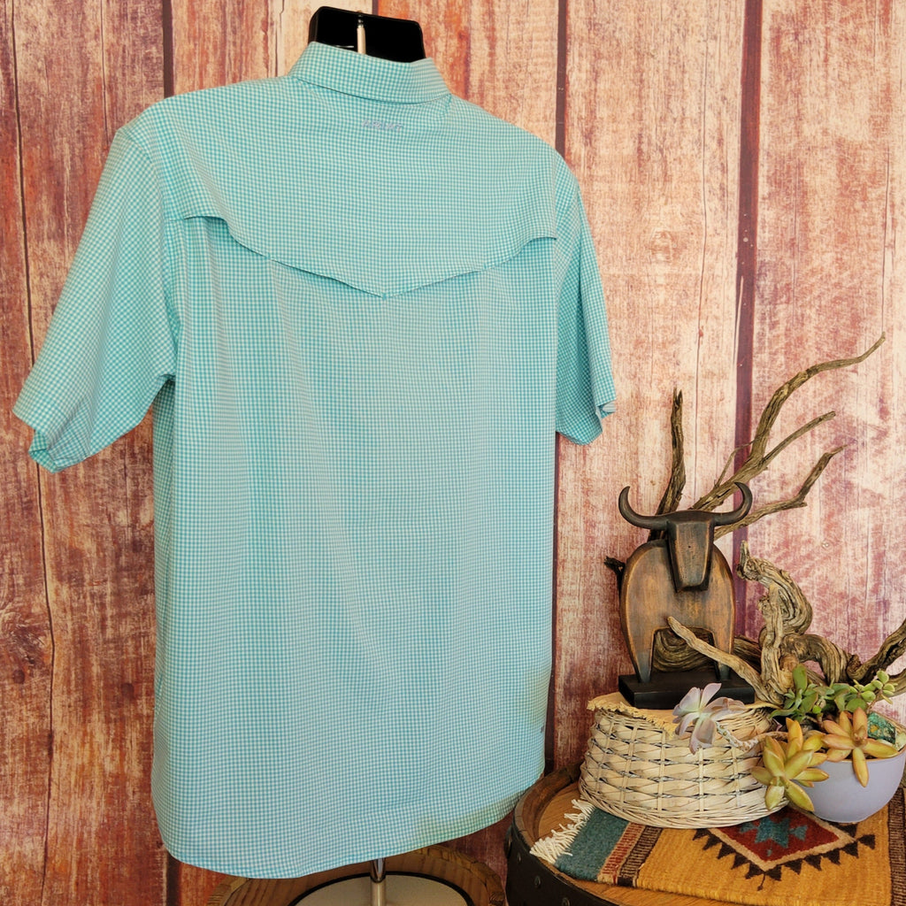 Men’s light turquoise checkered short-sleeve shirt by Ariat with snap buttons and two chest pockets, displayed on a mannequin against a rustic wooden wall. A barrel table nearby holds a basket and potted succulents, accented by driftwood, a bull skull sculpture, and a woven mat. Back view.
